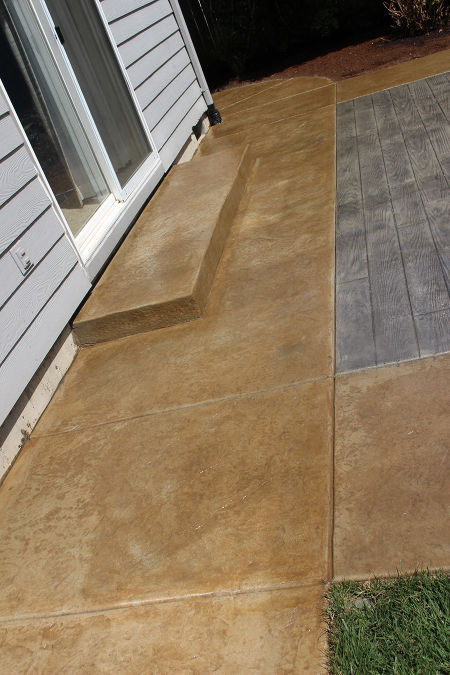 Brown concrete patio with a step near a white house and gray, wood-like patterned concrete.