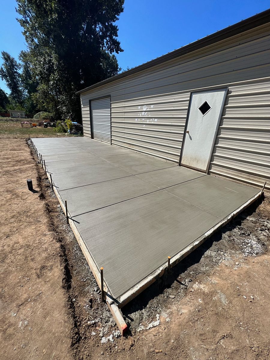 Freshly poured concrete patio next to a tan metal building with two garage doors and a white door.