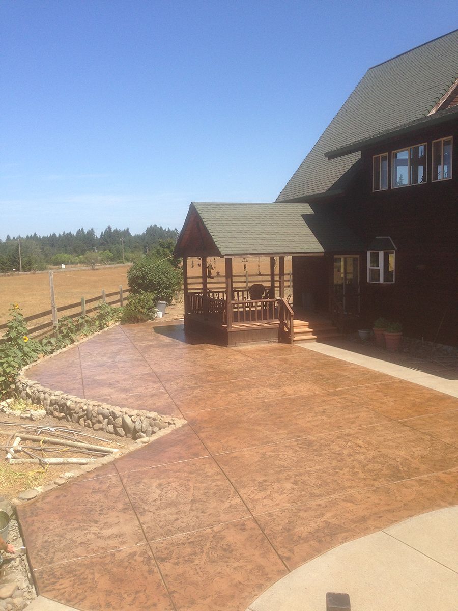 Brown stamped concrete patio with gazebo beside a dark brown house, farmland in the background.