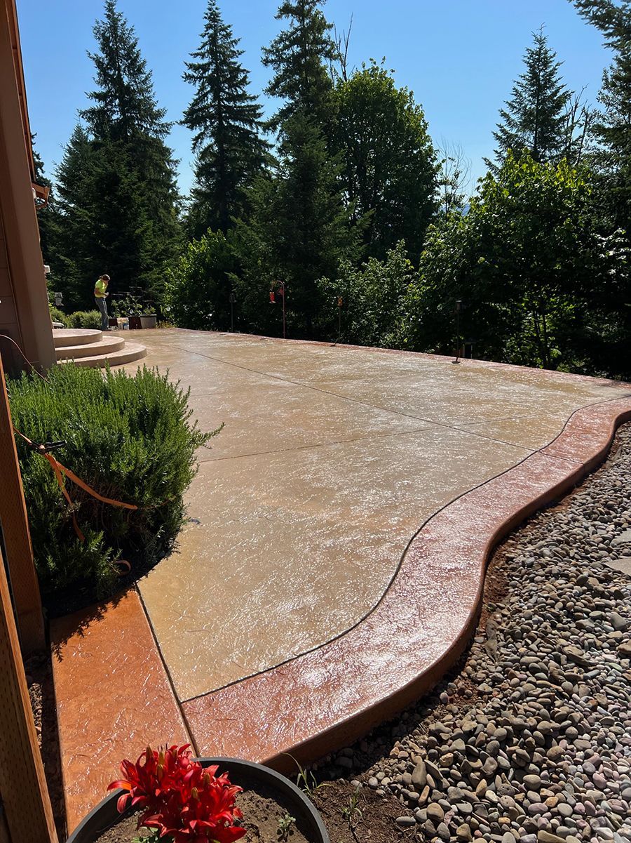 Stamped concrete patio with a curved edge, in shades of brown and orange, surrounded by trees and gravel.