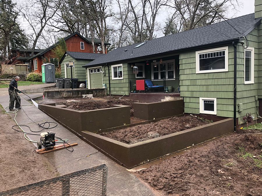Person pressure washing near raised garden beds, green house with dark roof.