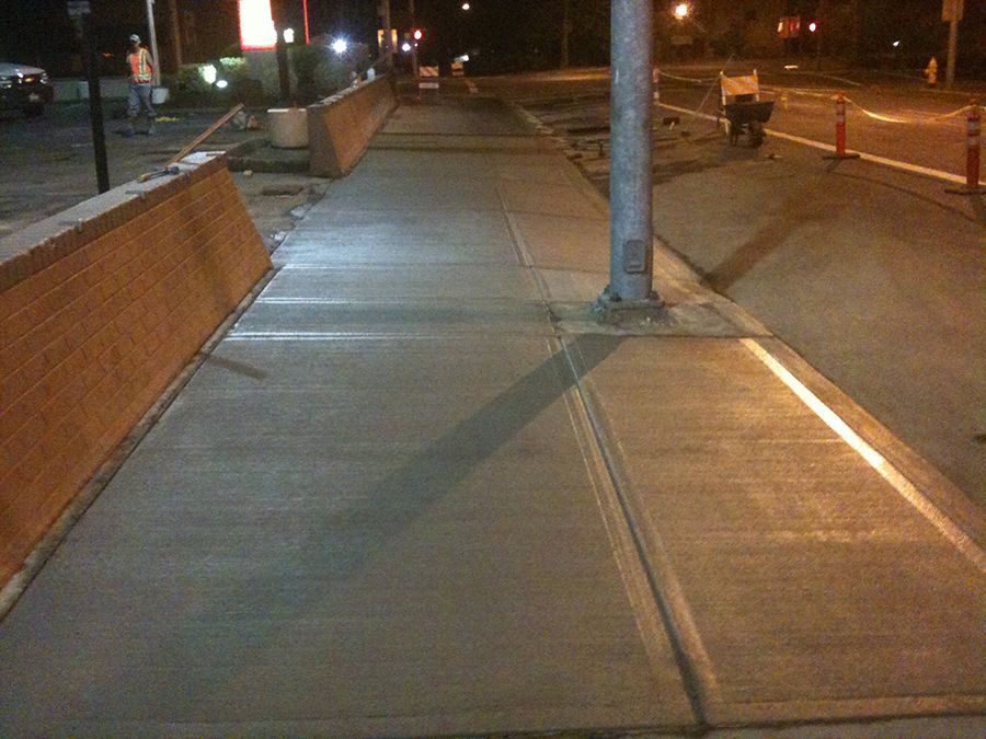 Newly poured concrete sidewalk with a light pole. Brick wall on the left, road on the right, dark setting.