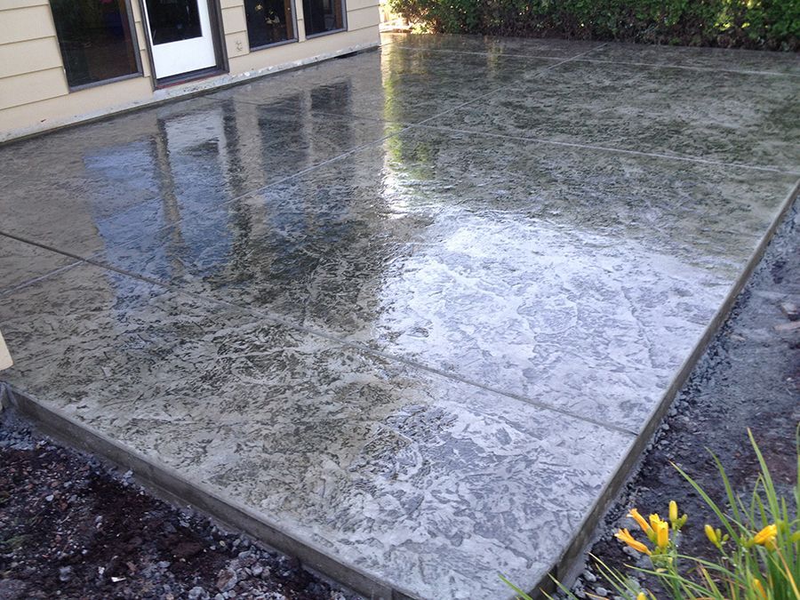 Concrete patio, textured finish, reflecting sky and adjacent house. Yellow flowers at the edge.