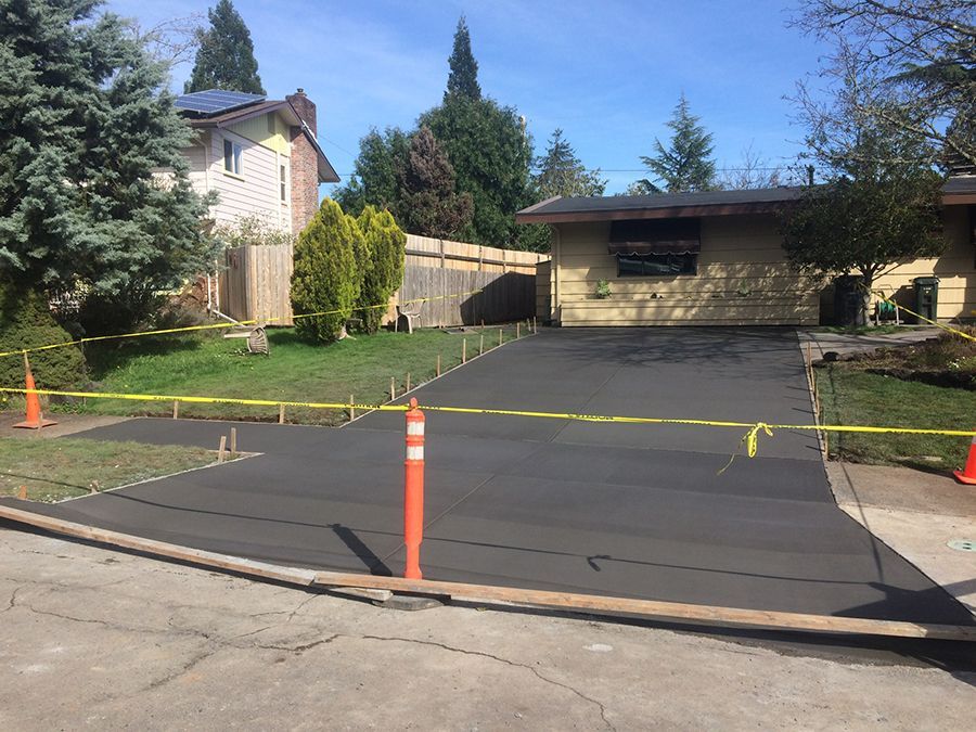 Freshly paved driveway with yellow caution tape, orange traffic cones, and a house in the background.