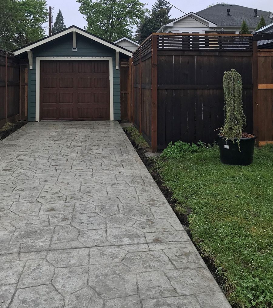 Driveway leading to a teal garage with a brown door, bordered by a dark wooden fence and a patch of grass.