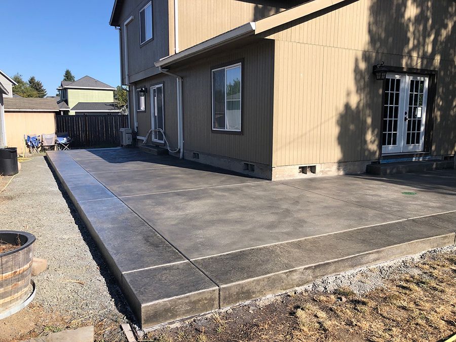 Concrete patio adjacent to a beige house with French doors, surrounded by gravel and grass, under a blue sky.
