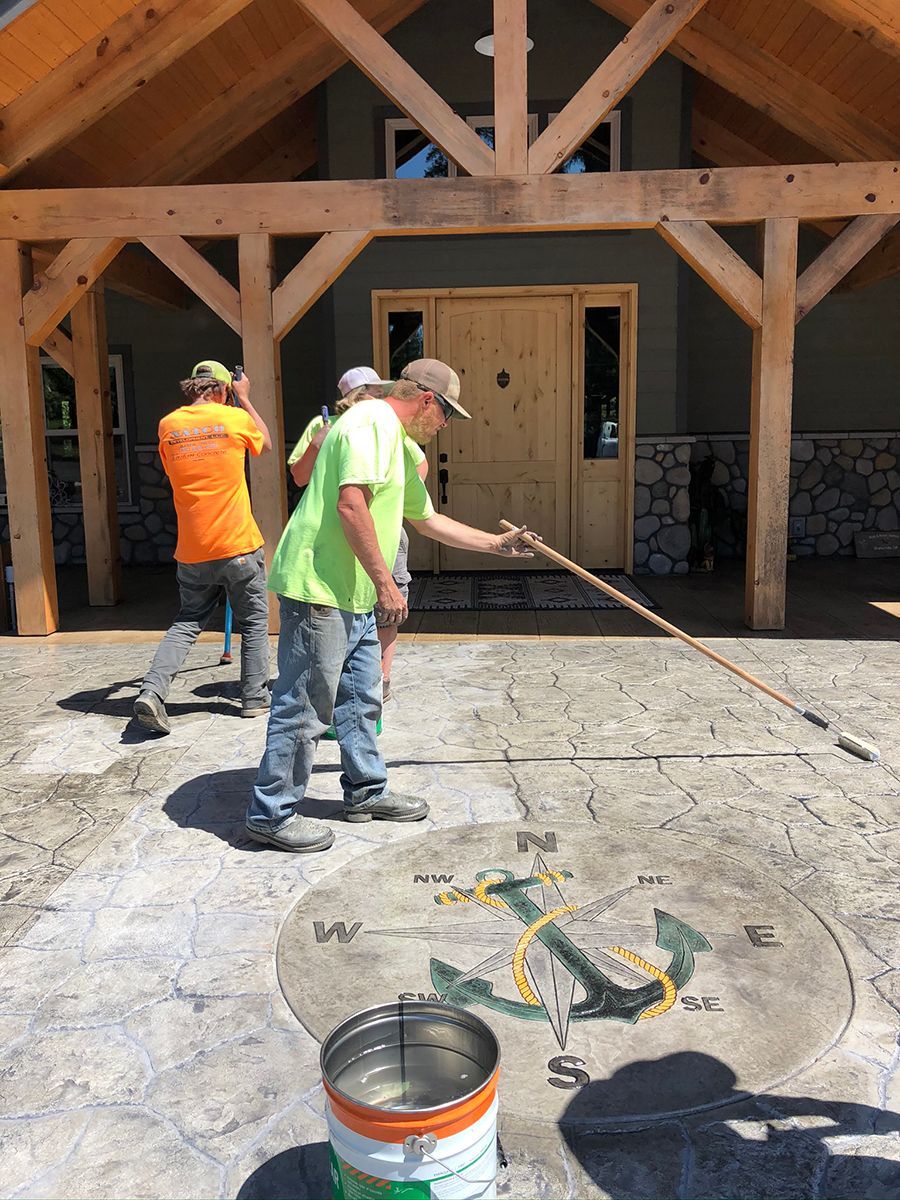 Workers applying color to a stamped concrete compass design on a patio. Wooden building in the background.