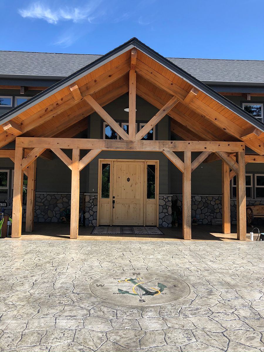 Wooden timber frame porch over a double door entrance with stone and gray siding, on a stamped concrete driveway.