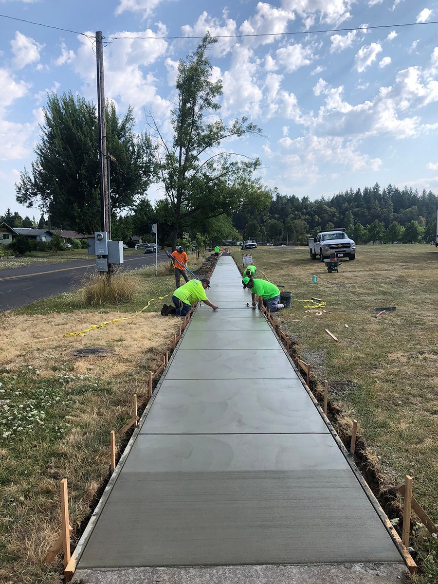 Workers smoothing fresh concrete sidewalk. Green shirts, tools, grass, road, blue sky.