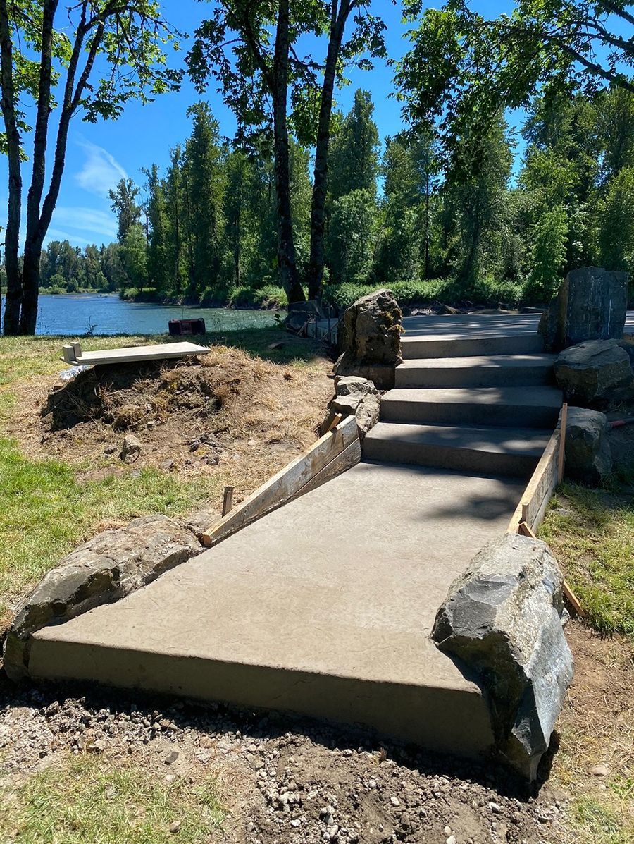Concrete path and stairs leading up a small hill near a body of water, bordered by rocks and grass.