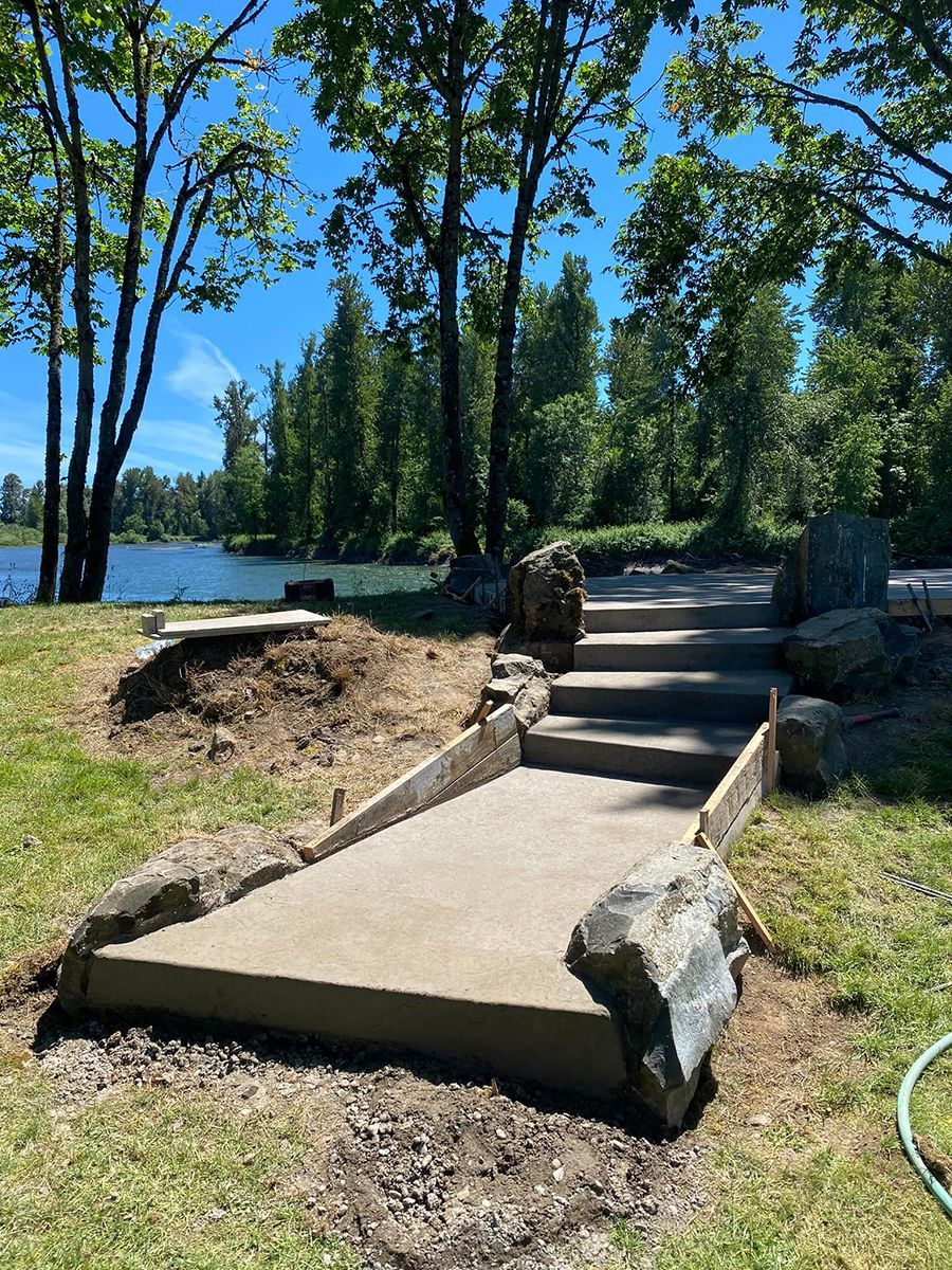 Concrete path and stairs leading towards a lake, framed by large rocks and grass, under a blue sky.