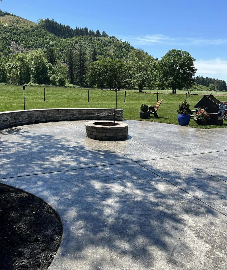 Stone patio with fire pit, stone wall, and chairs overlooking a grassy field with trees and a mountain.