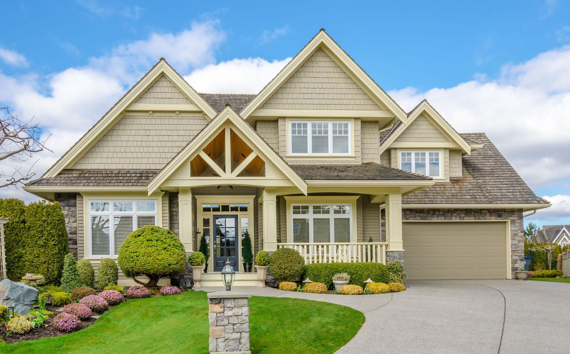 Tan and stone-colored house with a curving driveway, green lawn, and blue sky.