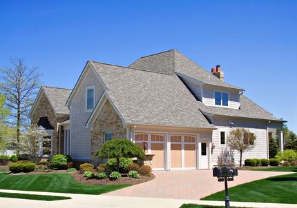 Two-story house with tan siding, gabled roof, and attached garage. Lush landscaping and blue sky.