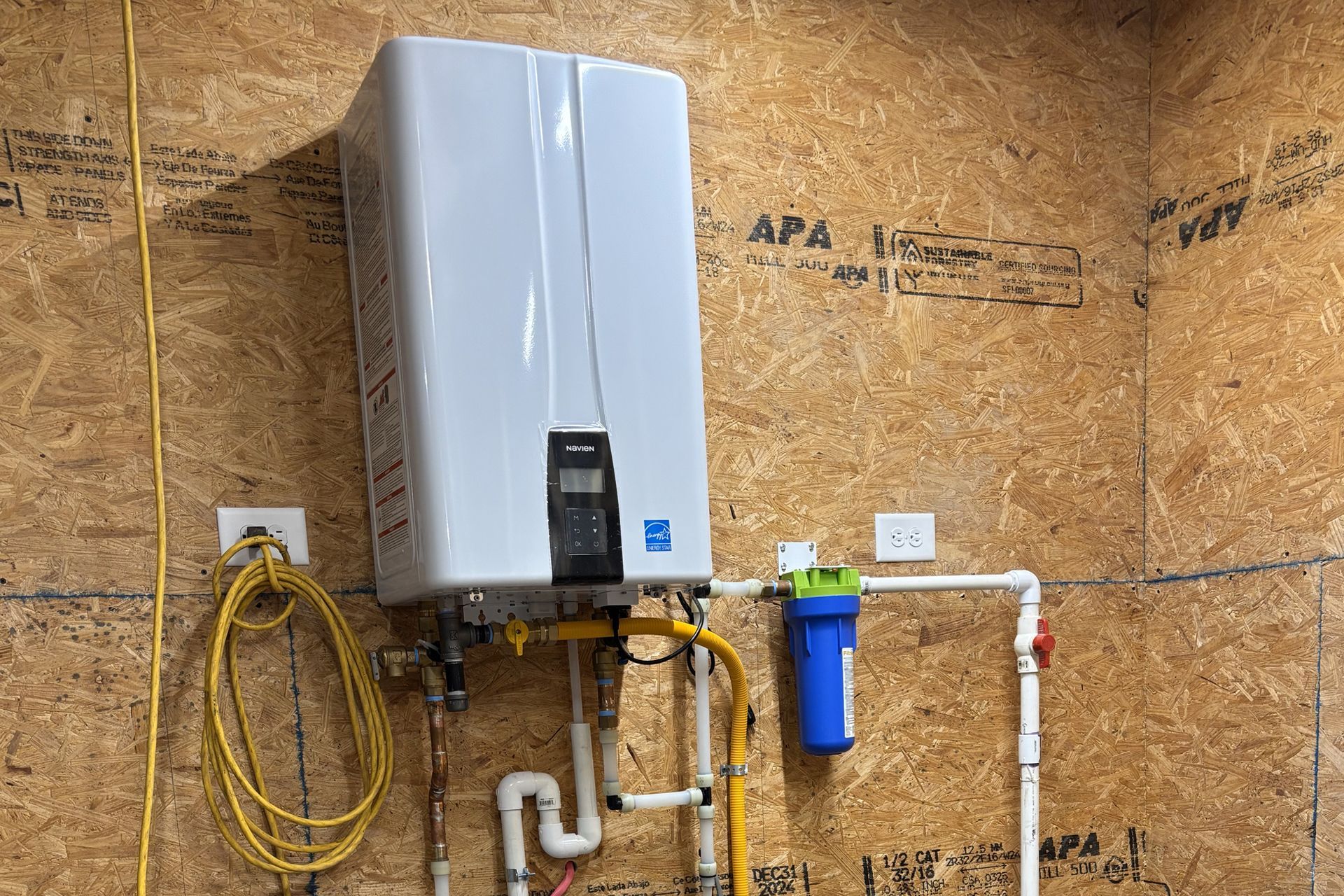 White tankless water heater mounted on a wood wall, with connected pipes and water filter.