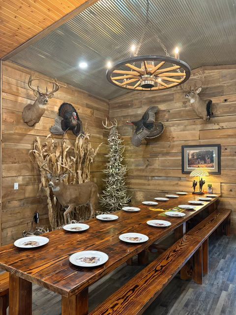 Rustic dining room with long wooden table, benches, and taxidermied animals on wood-paneled walls.