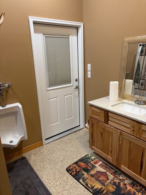 Bathroom with a white door, urinal, vanity, brown walls, and speckled flooring.