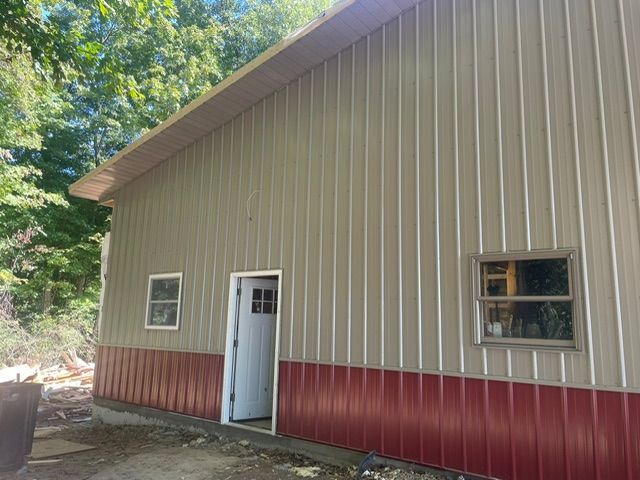 Tan and red metal-sided building with white trim, two windows, and a doorway set in a wooded area.