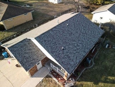 Overhead view of a house with a dark gray shingled roof, a driveway, and surrounding houses.