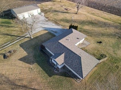 Aerial view of a gray-roofed house and a detached garage on a grassy hill; long driveway.