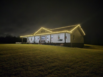 House lit with Christmas lights at night, visible on a grassy hill.
