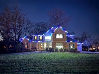 Two-story house at night with blue and white Christmas lights outlining roof and windows; green lawn.