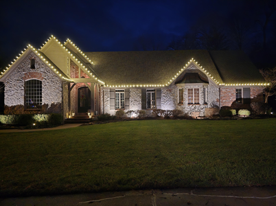 Brick house at night, lit with string lights along the roofline, illuminated lawn, and outdoor spotlights.