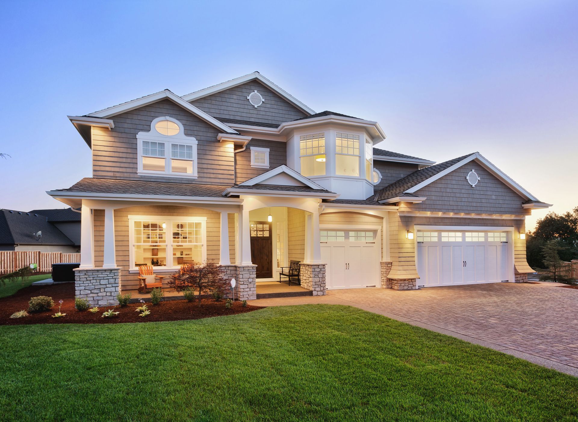 Two-story house with gray siding, white trim, and a stone entryway and garage, set in a green yard at dusk.
