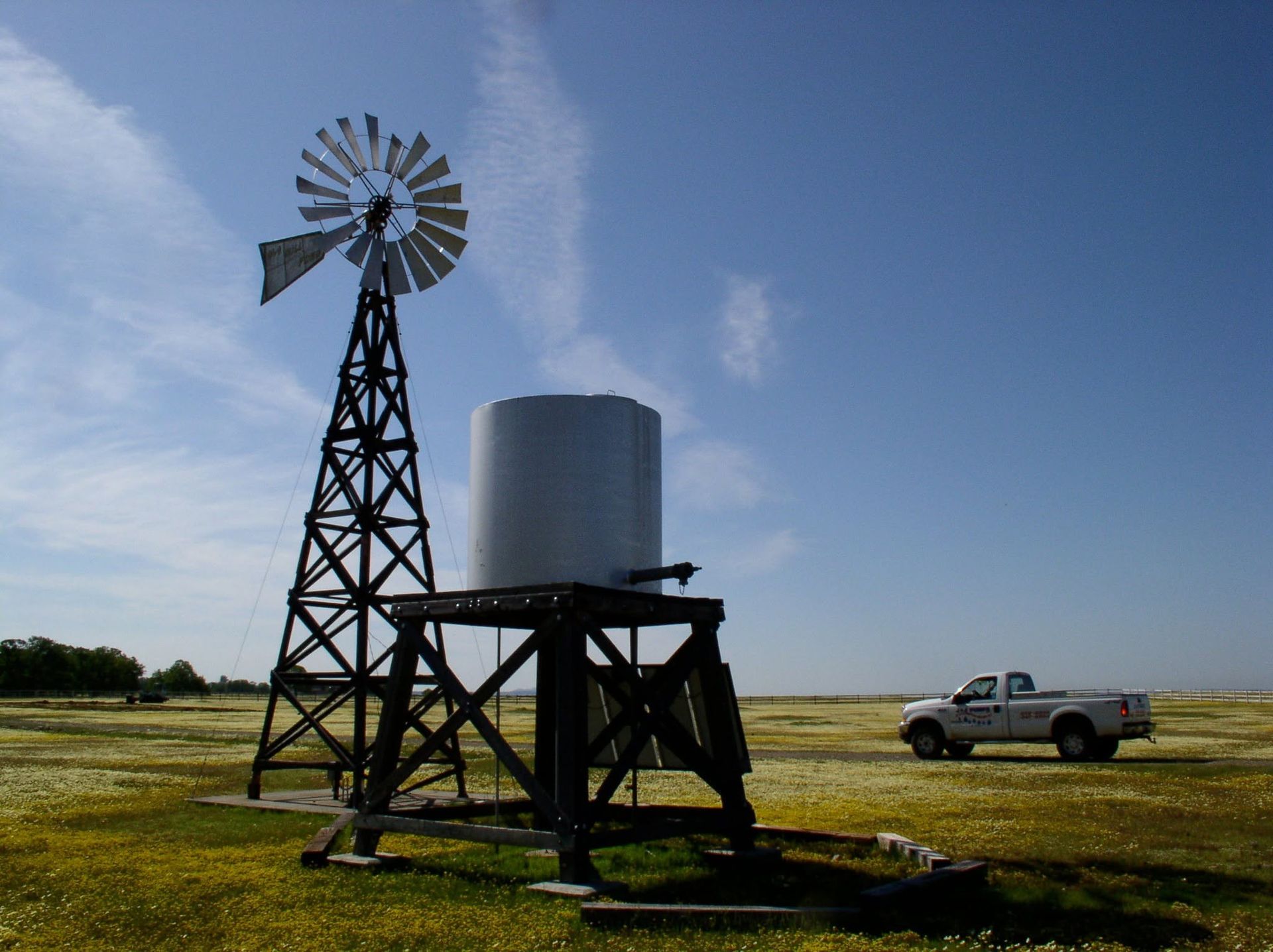 Windmill and water tank on a rural field, with a pickup truck under a clear blue sky.