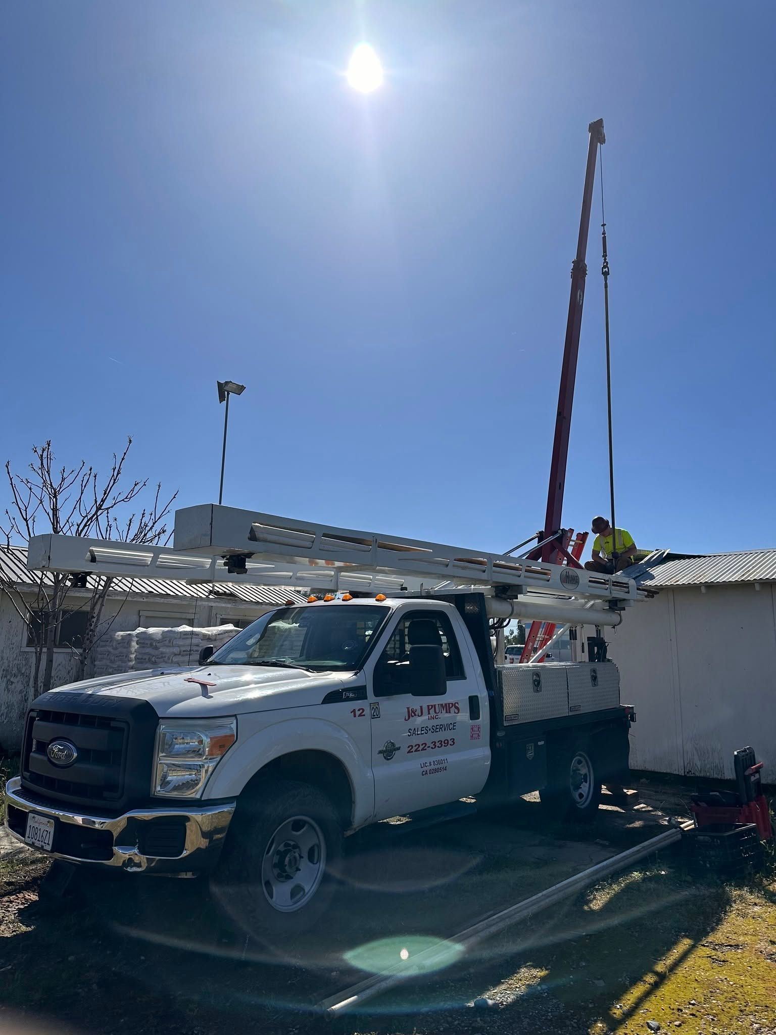 White utility truck parked by a building with roof work equipment and tall poles under bright sun