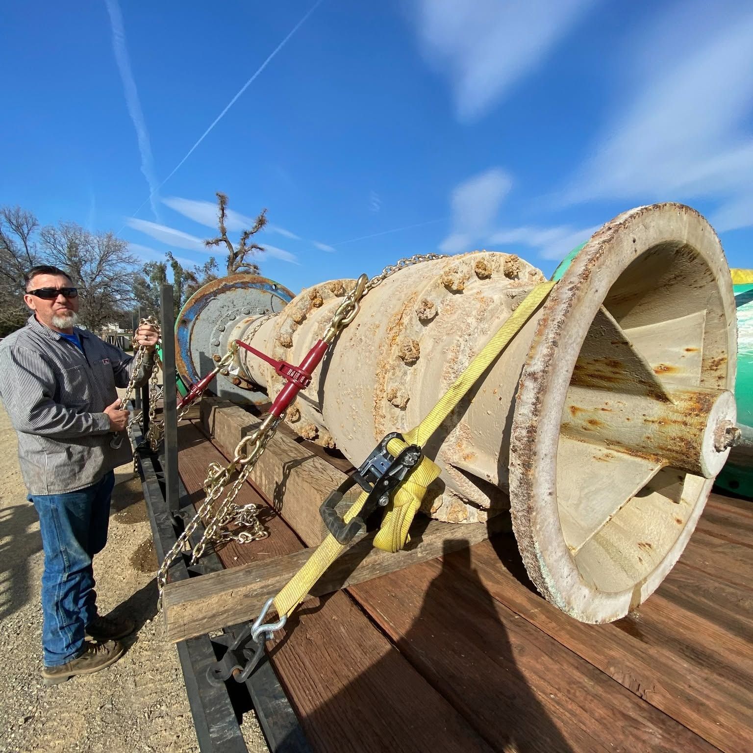 Large old metal turbine or pipe section strapped on a trailer, with a man standing beside it under a blue sky.