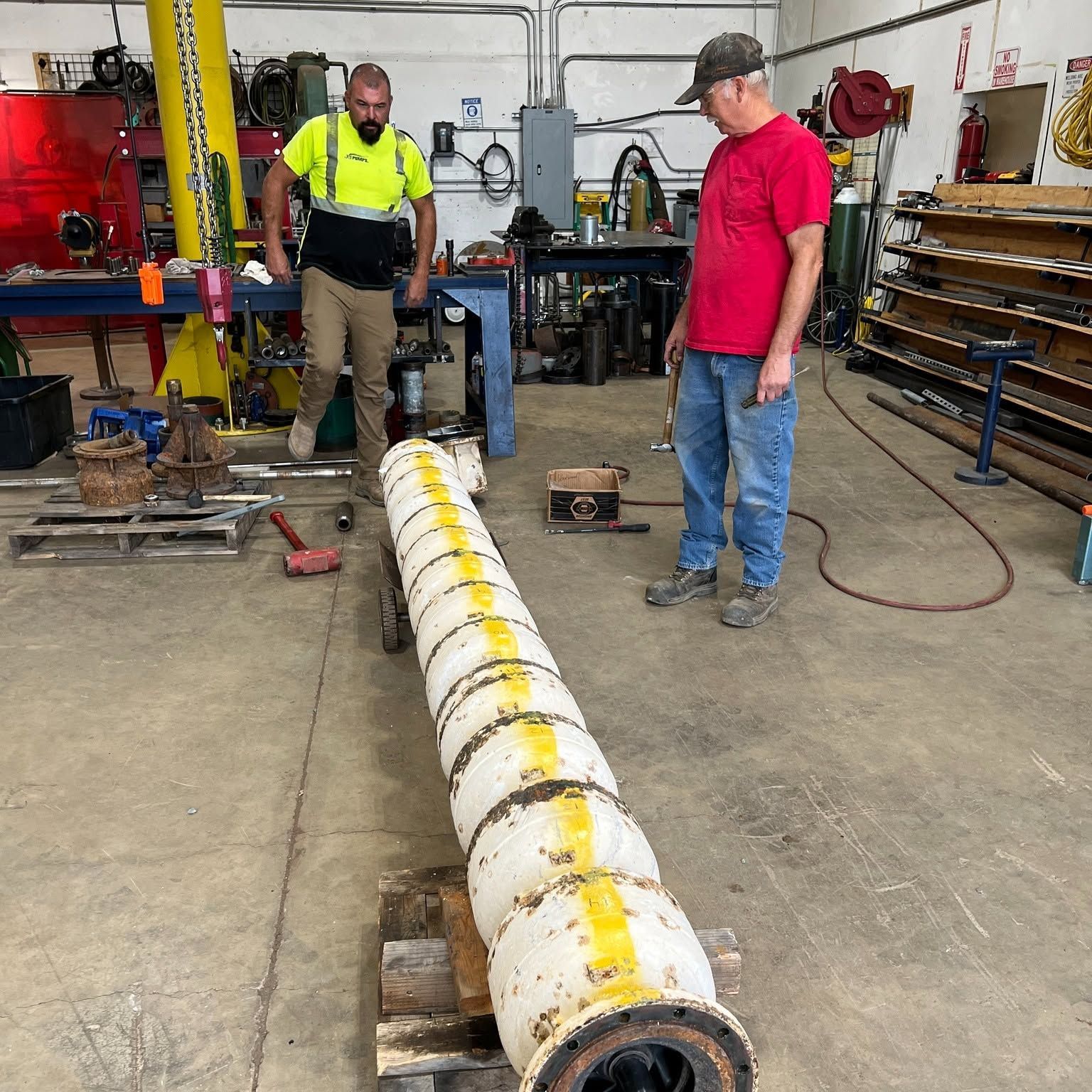 Two workers in a workshop inspect a long yellow-and-white cylindrical part on the floor.