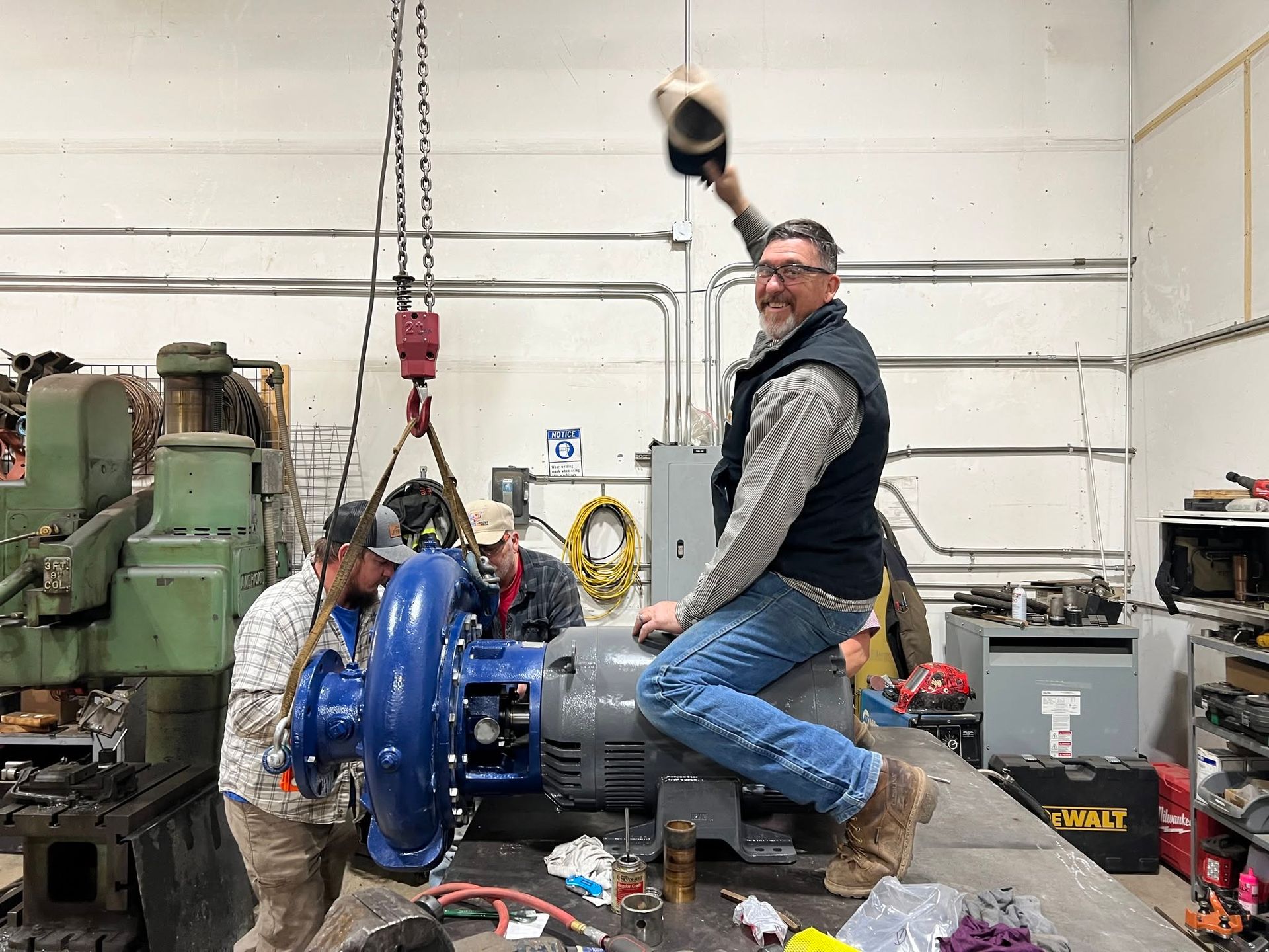 Worker kneeling on a factory bench beside a large blue mechanical part in a workshop