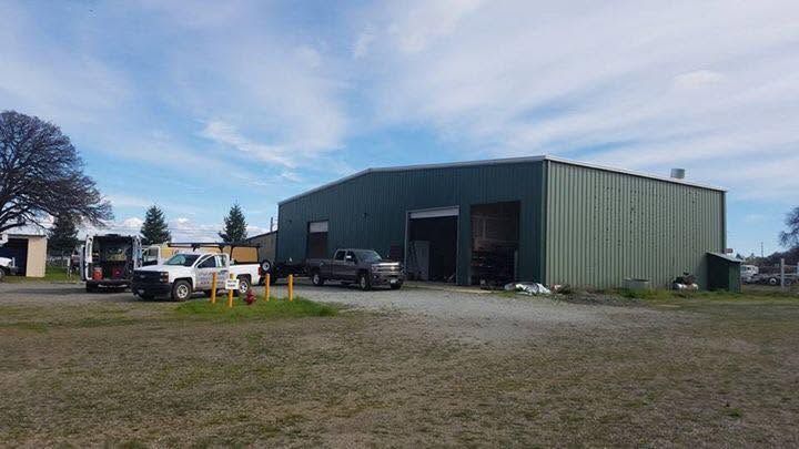Large green metal warehouse with open bay doors, parked vehicles, and a gravel lot under a cloudy sky