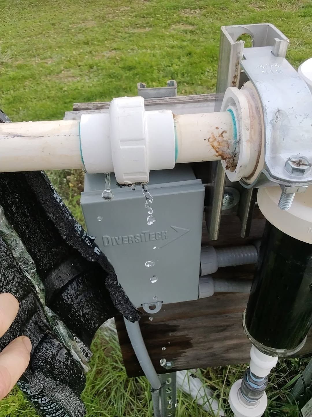 Close-up of white PVC pool plumbing and valve mounted beside a gray control box outdoors.