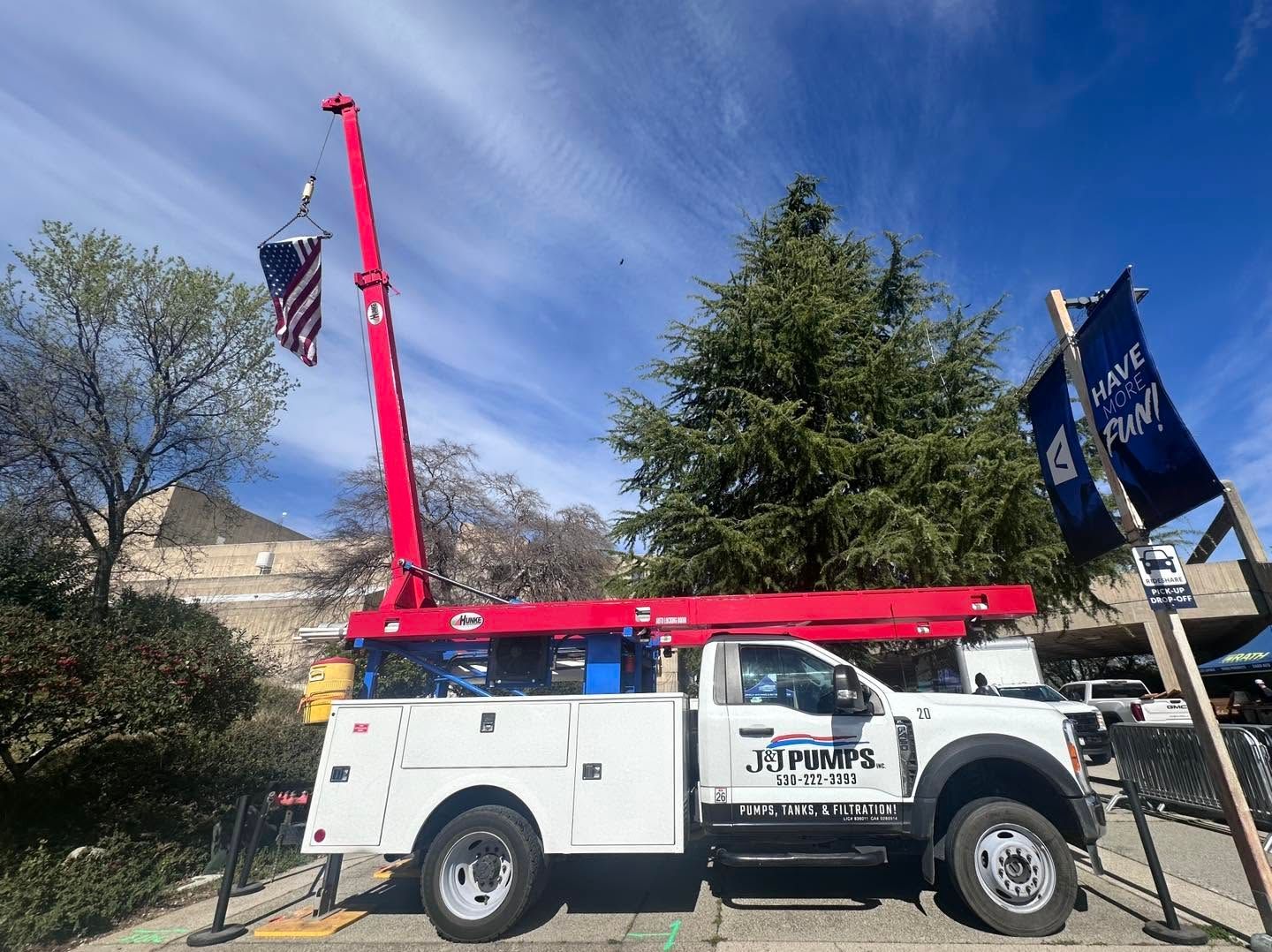 White utility truck with red crane arm parked outdoors near flags and trees under a blue sky.