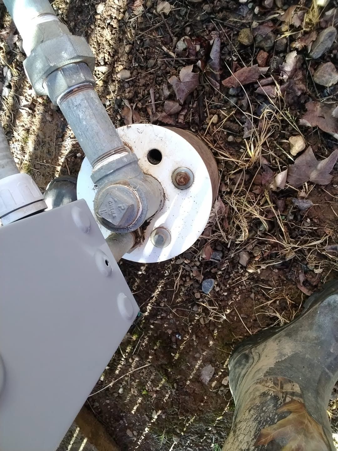 Utility pipes and valve on a white circular base outdoors beside soil and a boot