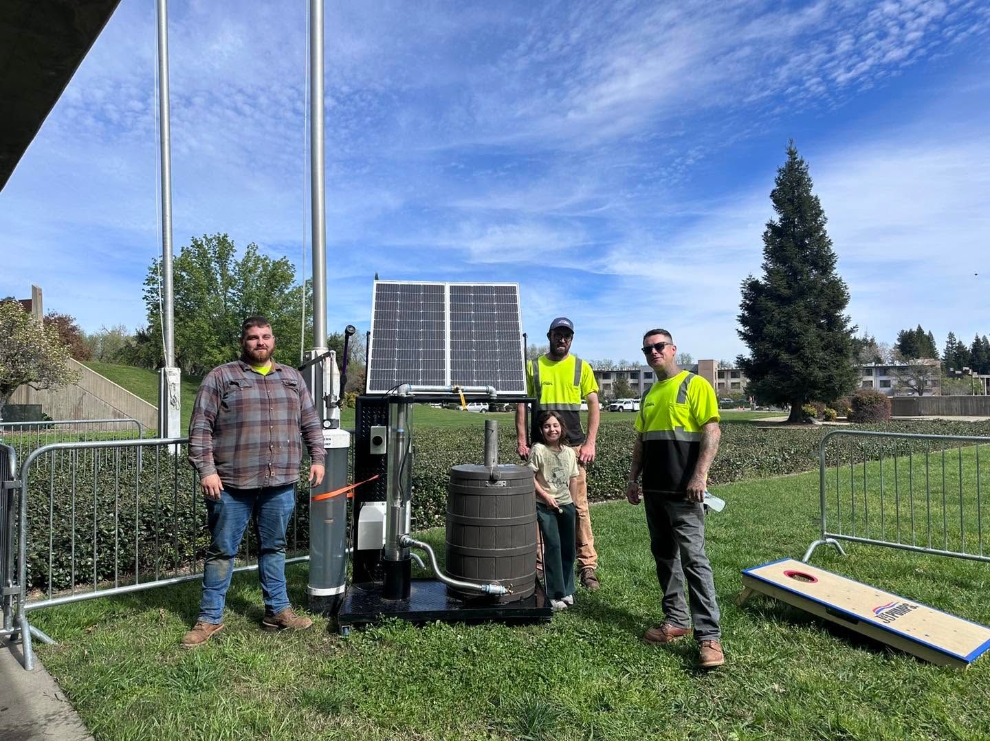 Three people stand beside solar equipment and panels on a grassy field under a blue sky.