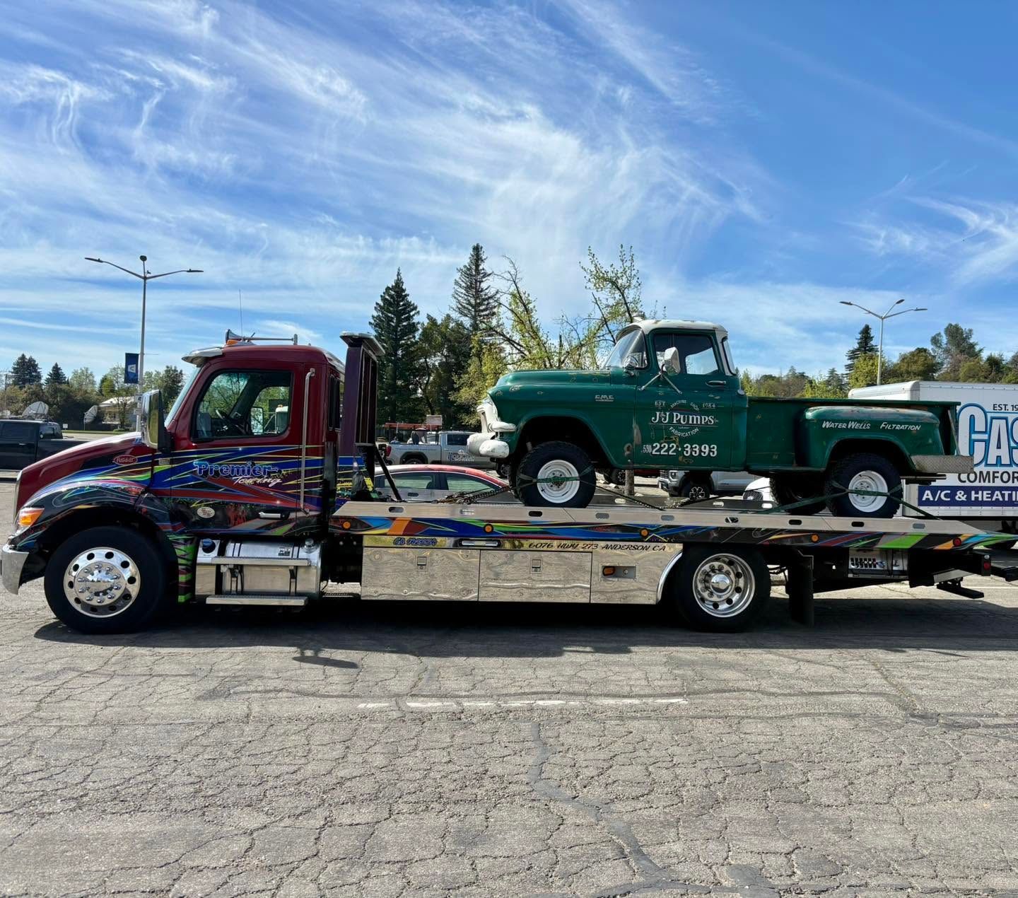 Red tow truck hauling a green vintage pickup on a flatbed under a blue sky.