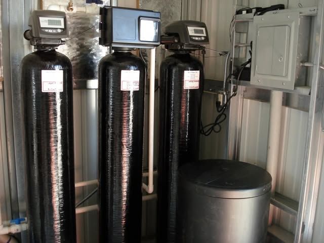 Water filtration equipment: three tall black tanks and a gray storage tank in a utility room.