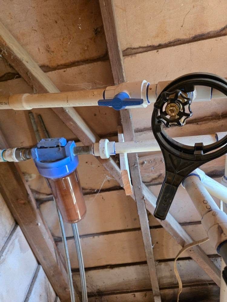 Plumbing pipes and blue water filter under a wooden ceiling, with a black steering wheel at right.
