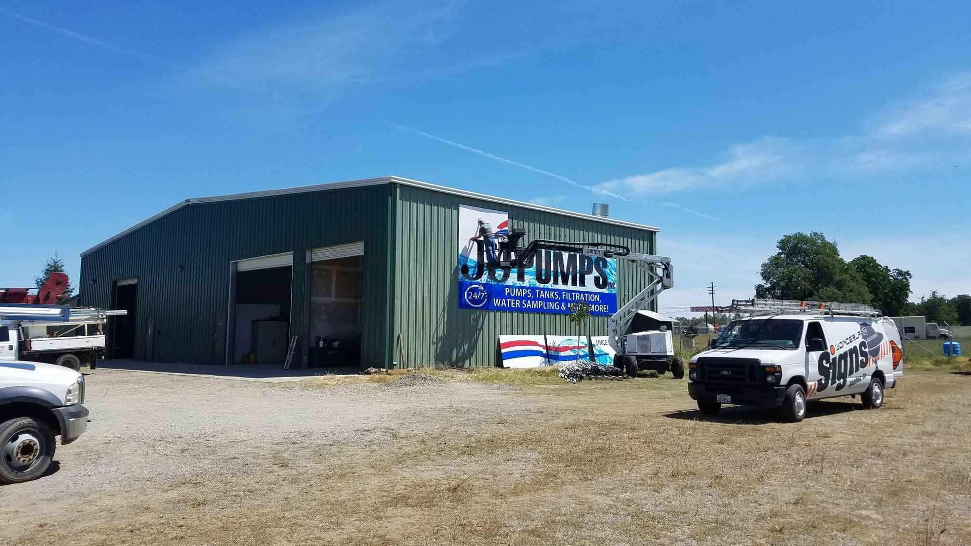 Green metal building with a large “PUMP” sign, parked white trucks, and an open gravel lot under a blue sky