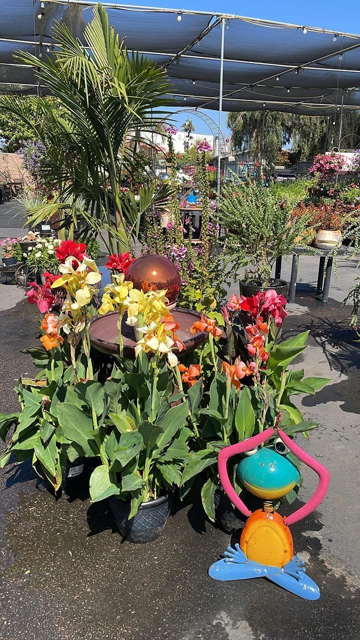 Colorful flowers and a metal sculpture in a nursery setting with shade overhead.
