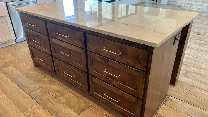 A wooden kitchen island with a stone countertop and multiple drawers, set against a light wood-plank floor.
