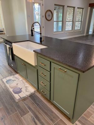 An olive green kitchen island with a white farmhouse sink, dark countertop, and a pumpkin-themed rug on a wood-look floor.