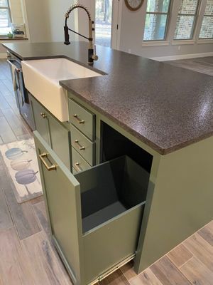 Sage green kitchen island with a farmhouse sink, dark countertops, and a pull-out trash drawer in an open-concept kitchen.