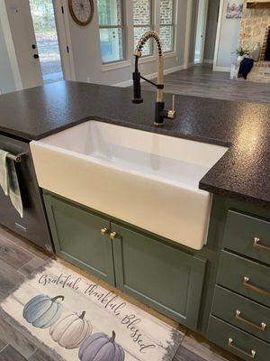 A white farmhouse sink with a tall black faucet in a kitchen with green cabinets and a pumpkin-themed floor mat.