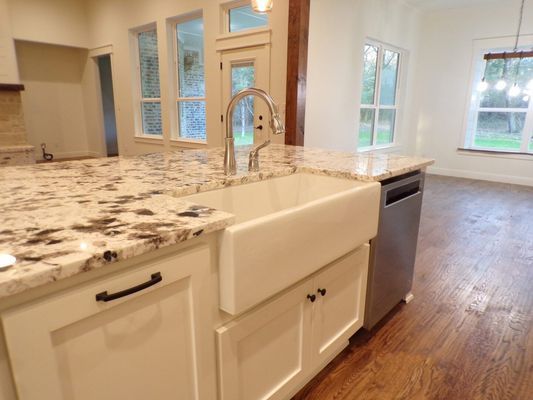 A farmhouse sink with a chrome faucet set in a white kitchen island with granite countertops and light-colored cabinets.
