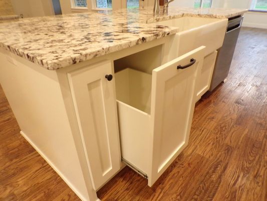 A white kitchen island with a pull-out cabinet containing a built-in trash bin, next to an apron-front sink and wood floors.