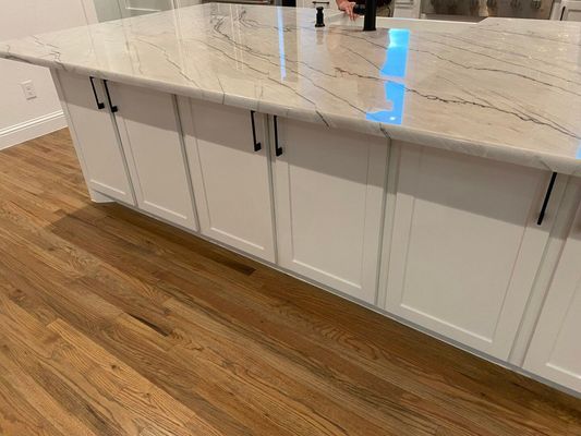 A kitchen island with white cabinets, dark hardware, and a marble countertop sits on light wood flooring.